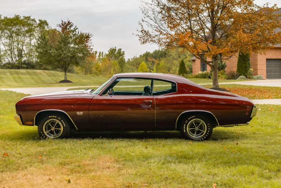 Vintage red muscle car parked on a grassy area with trees and a house in the background