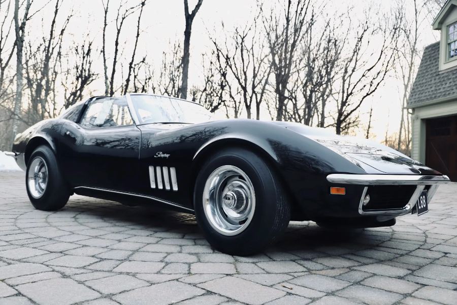 Black classic Corvette parked on a driveway with trees and a house in the background