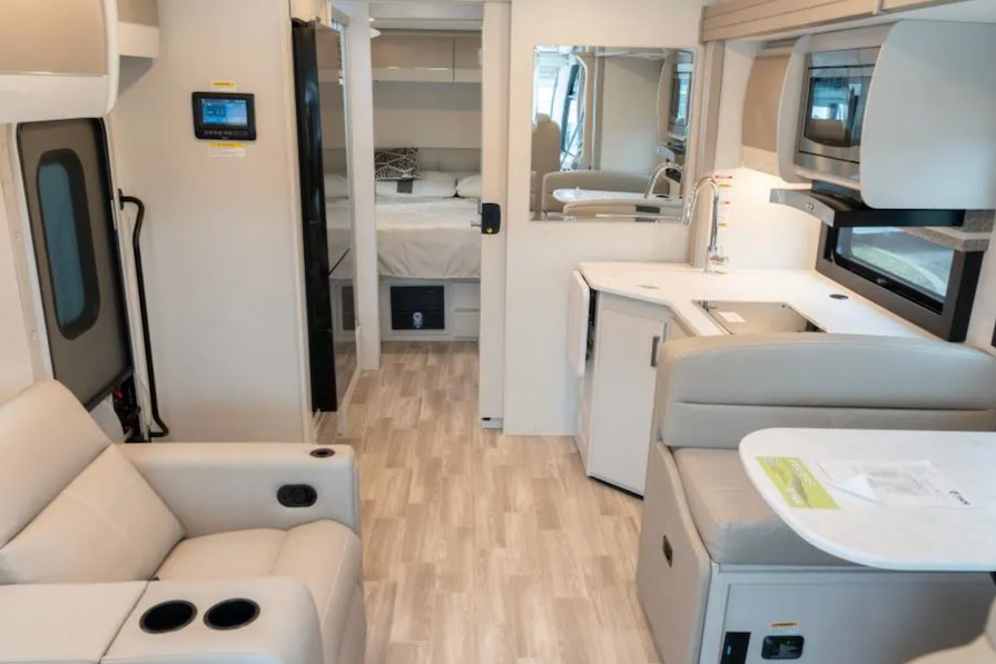 Interior of a modern RV with beige and gray furniture, including a couch, table, and kitchen area.
