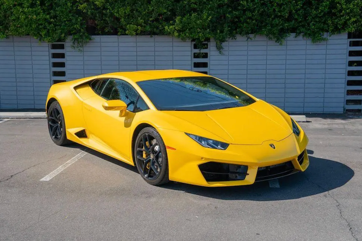 Yellow sports car parked in a lot with a blue wall and greenery in the background